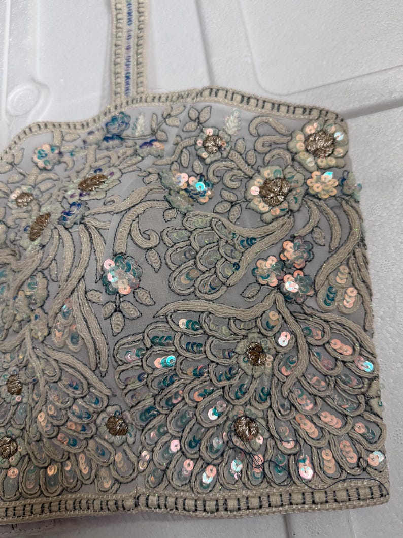 May include: A light grey, intricately embroidered top with floral and feather-like patterns. The design features sequins in shades of silver, blue, and gold. The edges are finished with a beaded trim, creating a detailed and elegant look.