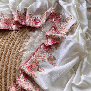 May include: A white fabric with a subtle crinkled texture featuring a scalloped edge embroidered with floral patterns in shades of pink, orange, and gold. The intricate embroidery includes detailed flower motifs and metallic thread accents.