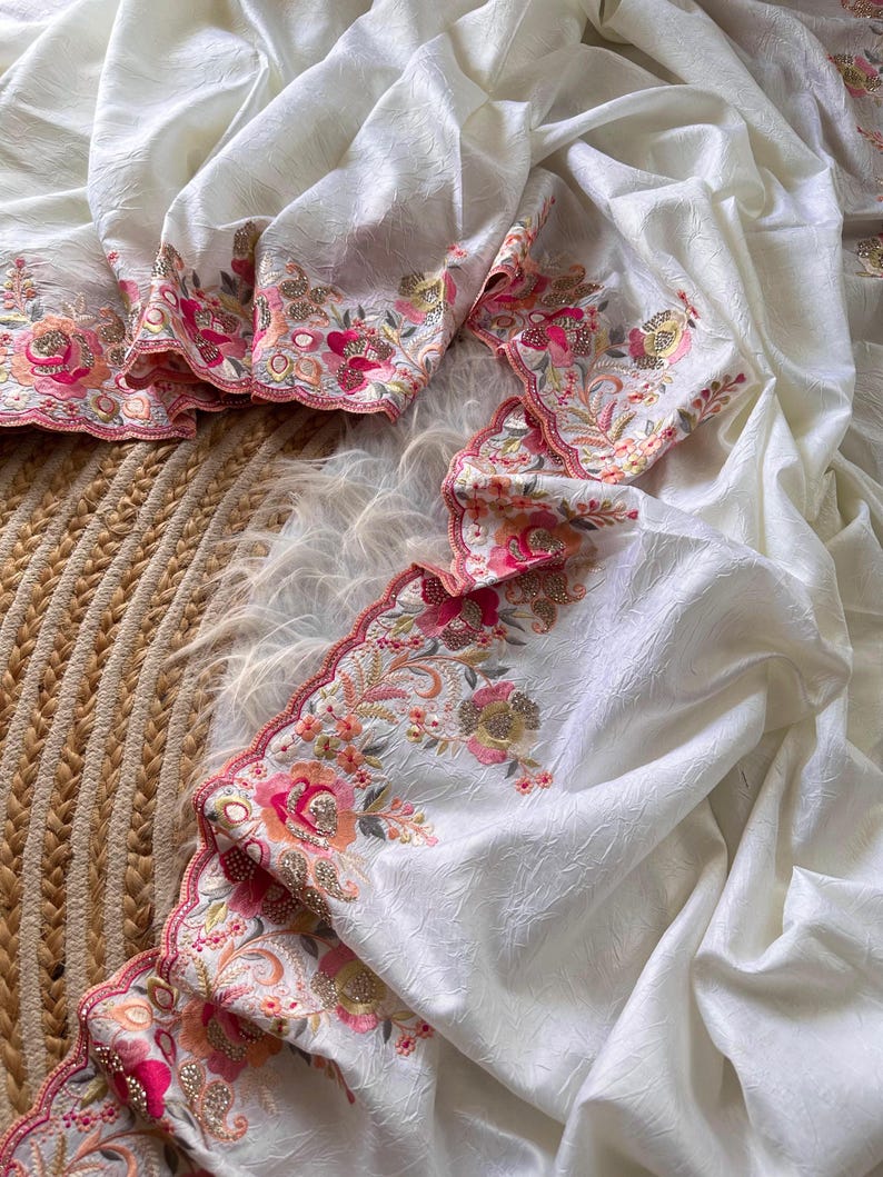 May include: A white fabric with a subtle crinkled texture featuring a scalloped edge embroidered with floral patterns in shades of pink, orange, and gold. The intricate embroidery includes detailed flower motifs and metallic thread accents.