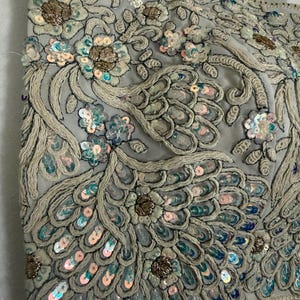 May include: Close-up of a light grey, heavily embellished top. The fabric is adorned with intricate embroidery, sequins, and beadwork in shades of silver, gold, and pastel blue. The design features floral and feather-like patterns.