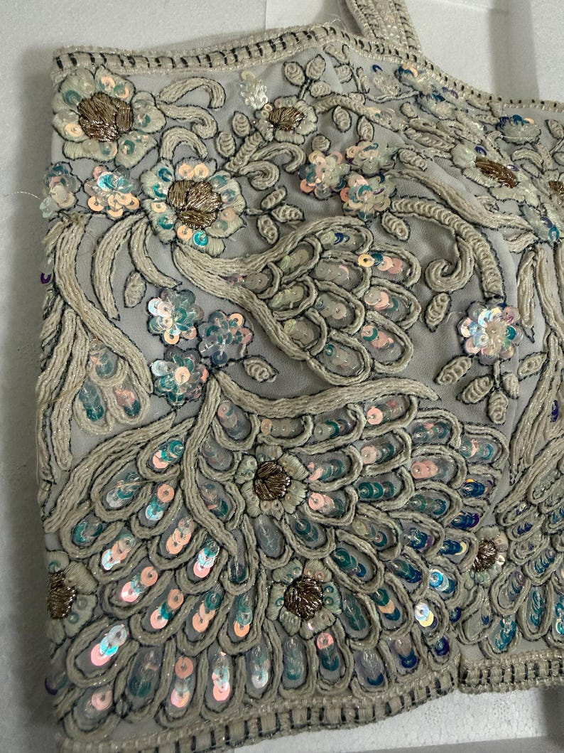 May include: Close-up of a light grey, heavily embellished top. The fabric is adorned with intricate embroidery, sequins, and beadwork in shades of silver, gold, and pastel blue. The design features floral and feather-like patterns.