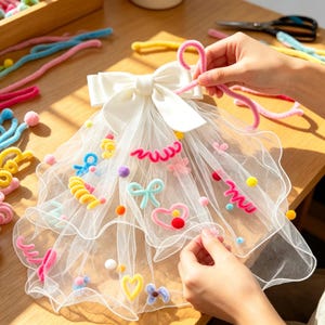 Op de afbeelding: Een witte tule sluier met een grote satijnen strik aan de bovenkant, versierd met diverse kleurrijke chenilledraad-vormen, waaronder hartjes, strikjes, spiralen en kleine pompons in roze, geel, blauw en paars verspreid over de stof.
