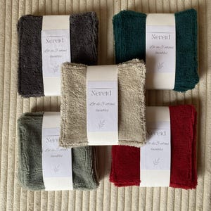 Peut inclure: Cinq ensembles de débarbouillettes réutilisables de couleurs variées : gris foncé, vert forêt, beige, vert olive et rouge. Chaque ensemble est emballé avec une bande blanche portant le nom de la marque "Nereid" et le texte "Lot de 5 cotons lavables".
