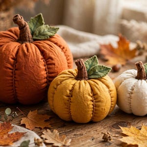 May include: Three fabric pumpkins in orange, mustard yellow, and cream. Each pumpkin features vertical stitched seams, a brown fabric stem, and two green felt leaves attached to the top. The pumpkins are arranged in a row on a wooden surface.