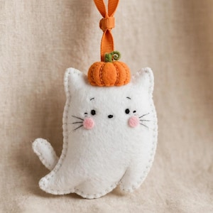 May include: A white felt cat ornament shaped like a ghost, featuring a small orange pumpkin on its head, pink cheeks, and black embroidered eyes and whiskers. An orange ribbon loop is attached to the top for hanging purposes.
