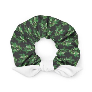May include: A fabric hair scrunchie featuring a dark gray background with a repeating pattern of green leaves and small white flowers. The accessory includes a solid white fabric bow tied at the bottom, adding a contrasting detail to the design.