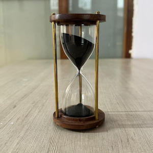 Könnte beinhalten: Eine Sanduhr mit Holzrahmen und Messingstützen. Die klare Glühbirne enthält schwarzen Sand. Die Sanduhr ist ein klassisches Zeitmessgerät, das oft zu dekorativen Zwecken oder zur Zeitmessung von Aktivitäten verwendet wird.