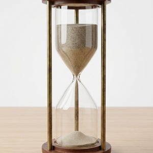 May include: An hourglass with a dark brown wooden frame and base, clear glass, and brass-colored metal supports. The hourglass contains tan sand. The hourglass is sitting on a light brown wooden surface.