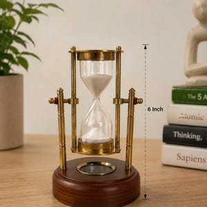 May include: An hourglass with a wooden base and brass-coloured metal frame. The glass bulb contains white sand. The hourglass is 15 cm tall. The base is a dark brown colour.