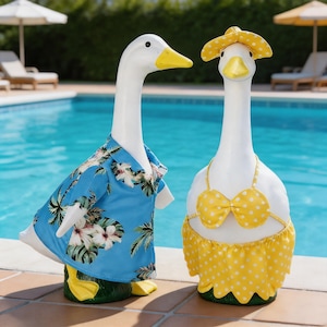 May include: Two white ceramic duck figurines, one in a blue Hawaiian shirt with a floral print, the other in a yellow polka dot bikini and matching sun hat. The ducks are near a swimming pool.