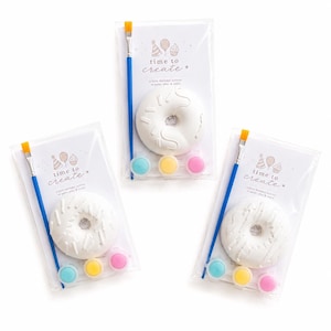 May include: Three craft kits in clear plastic packaging, each containing a white ceramic doughnut, a blue paintbrush with orange bristles, and three small paint pots in light blue, yellow, and pink. The card reads time to create.