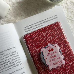 Könnte beinhalten: Eine rote Handyhülle mit weißem Tupfenmuster und einem dekorativen Aufkleber mit der Aufschrift "Buy me books & tell me I'm pretty". Die Hülle liegt auf einem offenen Buch.