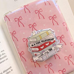 Può includere: Una custodia protettiva trasparente per e-reader o tablet con un motivo ripetuto di fiocchi rosa su sfondo rosa chiaro. Al centro è fissato un ciondolo in acrilico a forma di pila di libri con il testo IN A TOXIC RELATIONSHIP with my TBR.
