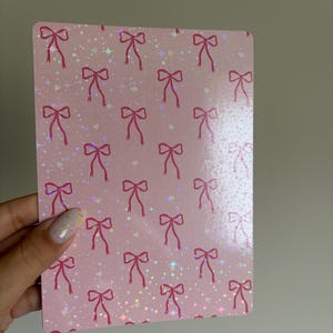 Peut inclure: Une carte rectangulaire rose avec un motif répété de nœuds rouges et de paillettes irisées. La carte est tenue dans une main avec un ongle peint.