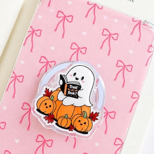 Peut inclure: Un support de téléphone transparent orné d'un fantôme de dessin animé assis sur des citrouilles orange, lisant un livre noir avec le texte "Treat or Book?" sur la couverture. Le support est fixé sur une coque rose pâle à motifs de nœuds.