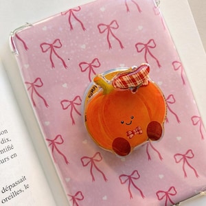 Puede incluir: Una funda protectora transparente para dispositivo móvil con un patrón repetitivo de lazos rosas y pequeños corazones blancos. En el centro hay un soporte circular para teléfono con forma de calabaza naranja con cara sonriente y un lazo a cuadros rojo.