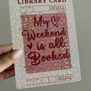 Peut inclure: Un autocollant rectangulaire en forme de carte de bibliothèque avec une finition holographique et scintillante. La carte comporte le texte "LIBRARY CARD" en haut, avec les mots "My Weekend is all Booked" en cursive. La carte a un fond rose.