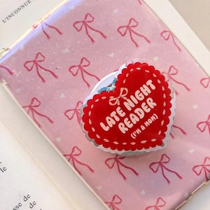 Peut inclure: Une coque de téléphone rose avec un motif répétitif de petits nœuds rouges et de cœurs blancs, accompagnée d'un support de téléphone en forme de cœur. Le support est rouge avec une bordure blanche festonnée et le texte LATE NIGHT READER (I'M A MOM).