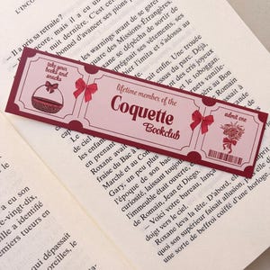 Könnte beinhalten: Ein rechteckiges Lesezeichen im Design eines Vintage-Tickets in Rosa- und Dunkelrottönen. Es enthält den Text "lifetime member of the Coquette Bookclub", "take your books and snacks" und "admit one" sowie Illustrationen eines Korbs und Blumen.