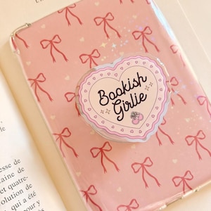 Puede incluir: Una funda de teléfono transparente con fondo rosa y un patrón repetitivo de lazos rojos y pequeños corazones blancos. En el centro, un agarre en forma de corazón con borde festoneado muestra el texto Bookish Girlie y dos cerezas.