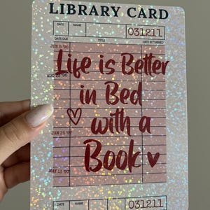 Könnte beinhalten: Ein holografischer Bibliothekskartenaufkleber mit dem Text "Life is Better in Bed with a Book". Das Kartendesign enthält Zeilen für Datum, Name und andere Bibliotheksinformationen. Der Aufkleber hat ein glitzerndes, irisierendes Finish.