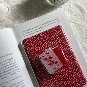 May include: A red and white polka-dotted phone case with a clear border, resting on an open book. A playing card with the Queen of Books is on top of the case. The text "Queen of Books" is visible on the card.