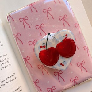 May include: A clear phone case with a pink background featuring a repeating pattern of red bows and small white hearts. Attached to the center is a heart-shaped phone grip with a blue and white gingham pattern and two glossy red cherries.