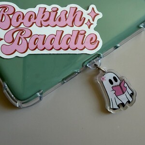 Puede incluir: Una funda transparente para teléfono con un inserto verde y una pegatina rosa y blanca que dice "Bookish Baddie". Un llavero con forma de fantasma con un libro está adjunto a la funda.