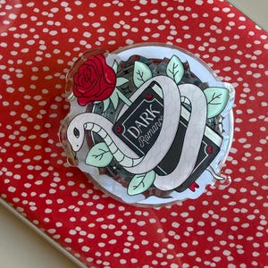 May include: A clear acrylic pin featuring a white snake coiled around two books with the words "Dark Romance". A red rose and green leaves surround the snake and books. The pin is on a red background with white polka dots.