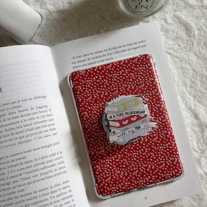 Peut inclure: Coque de téléphone rouge à pois blancs. Un autocollant sur la coque indique "IN A TOXIC RELATIONSHIP with my TBR" avec une pile de livres. La coque est posée sur un livre ouvert.