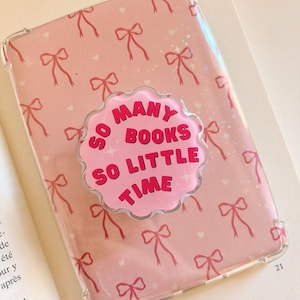 Op de afbeelding: Een doorzichtig beschermhoesje voor een mobiel apparaat met een lichtroze achtergrond en een herhalend patroon van rode strikjes en kleine witte hartjes. Een ronde grip met geschulpte rand bevat de tekst SO MANY BOOKS SO LITTLE TIME.