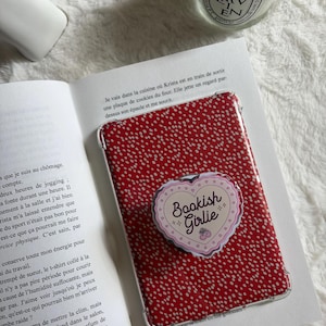 Peut inclure: Coque de téléphone rouge à pois blancs avec un pop socket en forme de cœur avec l'inscription "Bookish Girlie". La coque est posée sur un livre ouvert.