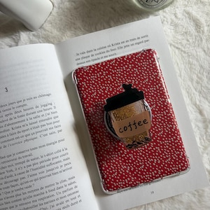Peut inclure: Un étui de téléphone rouge à pois blancs. Un motif de tasse à café avec le mot "coffee" est attaché à l'étui. L'étui est posé sur un livre ouvert.
