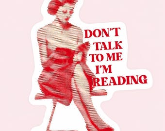 May include: Red and white sticker featuring a woman reading a book, with the text "DON'T TALK TO ME I'M READING". The image has a vintage, dot-matrix style, and is set against a light pink background.