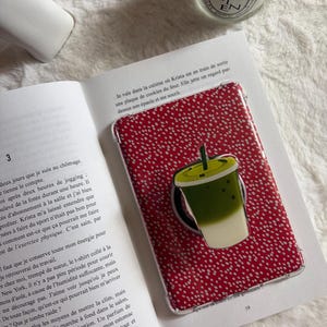 Peut inclure: Une coque de téléphone rouge à pois blancs et bordure transparente. Un pop socket vert et blanc représentant un matcha latte est fixé à la coque. La coque repose sur un livre ouvert.
