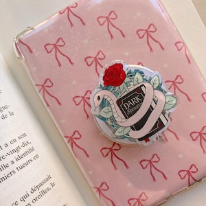 Peut inclure: Une coque de téléphone transparente sur fond rose avec un motif répétitif de petits nœuds rouges. Un accessoire circulaire en acrylique est fixé dessus, représentant un livre noir intitulé Dark Romance, entouré d'un serpent blanc et d'une rose rouge.