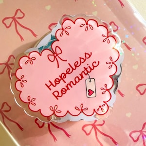 Peut inclure: Une broche en acrylique en forme de cœur avec un bord festonné, présentant un fond rose clair, des nœuds dessinés en rouge et le texte "Hopeless romantic" en écriture rouge. Une petite étiquette rectangulaire avec un cœur rouge est suspendue.