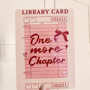 Peut inclure: Une carte de bibliothèque rectangulaire rose clair avec une finition holographique. La carte affiche le texte "LIBRARY CARD" en haut, "031211" dans les coins, et "One more Chapter" au centre avec un motif de nœud et de cœur.