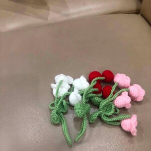 May include: Crocheted flowers in white, red, and pink with green stems and leaves. The handmade flowers are arranged on a neutral surface. The flowers are likely for decoration or crafting.