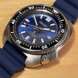 May include: A stainless steel Seiko dive watch with a blue dial and a black rotating bezel. The watch features white hour markers and hands, a date display, and a blue rubber strap. The watch is water resistant to 200 meters.