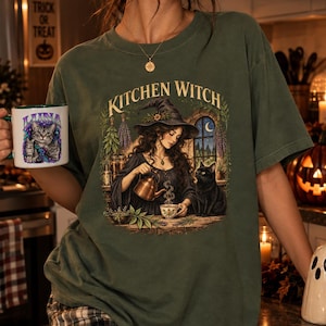 Op de afbeelding: Een donker olijfgroen t-shirt met korte mouwen en een afbeelding van een heks die thee inschenkt bij een zwarte kat. De tekst "KITCHEN WITCH" staat boven de illustratie. Een witte mok met een kat en de tekst "LUNA" wordt ernaast gehouden.