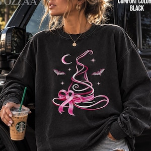 May include: A black long-sleeve crewneck sweatshirt featuring a graphic of a witch hat formed from a pink ribbon. The design includes a crescent moon, stars, and two bats. The text COMFORT COLORS BLACK is visible in the upper right corner of the image.