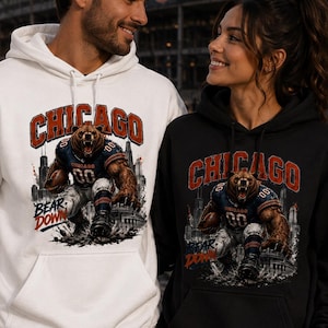 May include: A white hoodie and a black hoodie, each featuring a graphic print of a bear in a football uniform running with a ball. The text CHICAGO appears in large red letters above the bear, with the words BEAR DOWN printed in blue and red below.