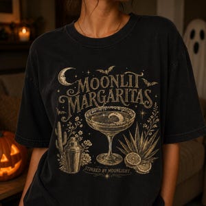 Può includere: Una t-shirt nera a maniche corte con una stampa grafica che riporta il testo "MOONLIT MARGARITAS" e "STIRRED BY MOONLIGHT". Il design include un bicchiere da margarita, uno shaker, un cactus, foglie di agave, fette di lime, pipistrelli e una luna crescente.