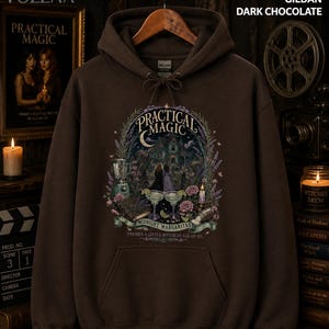 Op de afbeelding: Een donker chocoladebruine hoodie met een grafische print met de tekst Practical Magic, Midnight Margaritas en There's a little witch in all of us. Het ontwerp bevat een huis, twee figuren, margarita's en diverse florale elementen.