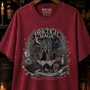 May include: A chili red short-sleeve t-shirt featuring a graphic print of a house, two figures, margaritas, and a blender. The text reads Practical Magic, Midnight Margaritas, and There's a Little Witch in All of Us. The shirt has a crew neckline.