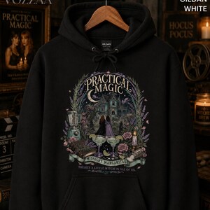 Op de afbeelding: Een zwarte hoodie met een grafische print met de tekst Practical Magic, Midnight Margaritas en There's a little witch in all of us. Het ontwerp bevat een huis, twee figuren, een blender, margarita's en bloemmotieven.