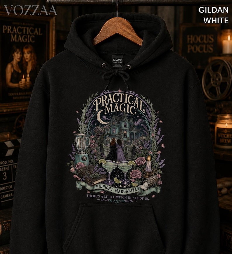 Op de afbeelding: Een zwarte hoodie met een grafische print met de tekst Practical Magic, Midnight Margaritas en There's a little witch in all of us. Het ontwerp bevat een huis, twee figuren, een blender, margarita's en bloemmotieven.
