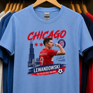 May include: A light blue t-shirt featuring a graphic of Robert Lewandowski flexing his arm. The design includes the text CHICAGO, LEWANDOWSKI, THE POLISH GOAL MACHINE, a Chicago skyline silhouette, three red stars, a soccer ball, and a signature.