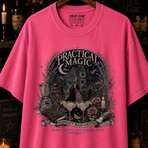 May include: A neon pink short-sleeve t-shirt featuring a graphic print of a house, two figures, a blender, and two margaritas. The text reads Practical Magic, Midnight Margaritas, and There's a Little Witch in All of Us. The shirt has a crew neckline.